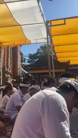 A group of people dressed in white are sitting under a yellow and white awning. The people appear to be at a religious ceremony. The architecture in the background suggests a temple. The sky is clear and blue. One person in the foreground is wearing a white shirt and has their head down. Another person is wearing a white head covering and a flower in their hair. The overall mood is serene and respectful.