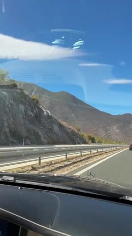 The video shows a view from inside a car looking out the windshield. The car is on a highway, with a mountain range on the left side of the road and other cars in the distance on the highway. The sky is blue with some white clouds. The dashboard of the car is visible in the foreground. The sun is shining and it appears to be daytime.