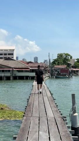 A person is walking away from the camera down a wooden plank pier over water. The person is wearing a black shirt, black shorts, white socks, and white sneakers. They also have a black cap and a black bag slung across their body. The pier is made of wooden planks, and the water is blue with a little green growth along the edge of the pier. On either side of the pier are buildings with red roofs, some of which are built on stilts over the water. In the background, there is a city skyline with several tall buildings and a blue sky with some white clouds.