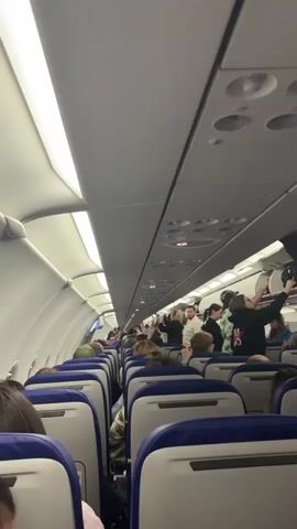 The video shows the interior of a commercial airplane cabin, viewed from the perspective of a passenger in a window or middle seat. The aisle is visible, lined with rows of seats on either side. Many passengers are seated, and some are standing in the aisle, either retrieving items from overhead compartments or interacting with others. Overhead bins are open, and luggage is being stowed. The lighting appears to be artificial, typical of an aircraft cabin. The camera is shaky, indicating movement. The overall scene depicts boarding or deplaning activity, or possibly a delay or unusual situation within the cabin.
