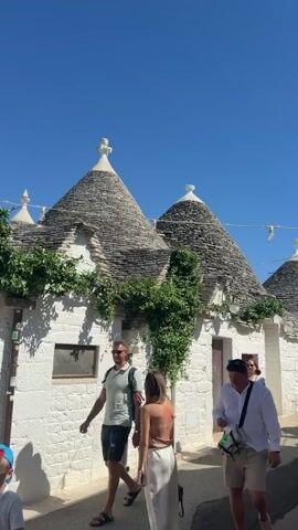 The video shows a street lined with traditional Trulli houses in Alberobello, Italy. The houses are whitewashed with conical stone roofs. Several people are walking down the street. On the left, a man in a green polo shirt and shorts walks away from the camera. To his right, a woman in a white outfit walks towards the camera, carrying a black bag. Behind her, a man in a white shirt and beige shorts also walks towards the camera. Further down the street, another man and woman are visible. The sky is clear and blue.