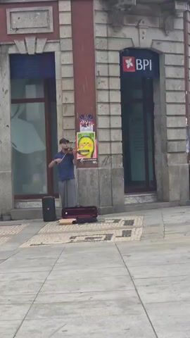 A person wearing a blue shirt and gray pants plays the violin on a sidewalk in front of a building. A black speaker and a red violin case are on the ground nearby. Two people, a woman and a man wearing a white baseball cap, walk past in the foreground. The building has a sign that reads "BPI" and large windows. The sidewalk is paved with stone. The video captures the street musician performing and pedestrians passing by.