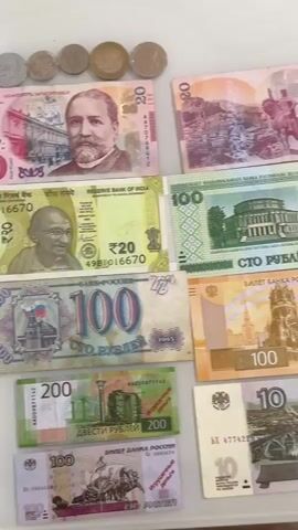 The video displays a collection of various banknotes and coins spread across a white surface. The currencies appear to be from different countries, including Russia, India, and possibly Ukraine, along with US dollars. The banknotes include denominations such as 100, 200, and 1000 rubles, 20 and 500 rupees, a 1 dollar bill, and others that are less distinct. There are also several coins at the top left of the frame. The lighting suggests an indoor setting, possibly a table, with some reflections on the surface. The camera moves smoothly, providing a top-down view of the currency display. No individuals are visible in the video.