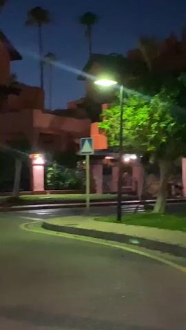 The video shows a nighttime scene on a street with palm trees and a building in the background. The building appears to be made of light-colored stone and has a warm glow from interior lights. Several light posts illuminate the area. Two women, dressed in black dresses, are walking away from the camera down a curved road. The woman on the right has blonde hair and is carrying a small purse. The woman on the left has dark hair and is also carrying a purse. They are walking on a paved road with yellow and white markings. The environment is a resort or residential area with manicured lawns.