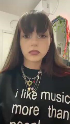 A young woman is holding a hair straightener in her hands, straightening her bangs. She is wearing a black, long-sleeved shirt that says "I like music more than people." She has long brown hair and is wearing multiple necklaces and rings. Behind her is a mirror decorated with lights and vines, a white door, a clock, and an air conditioner.