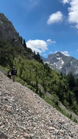 Two individuals are hiking up a steep, rocky mountain trail. The person in front is wearing a dark backpack, and the person behind them is wearing a lighter backpack. The trail is covered in loose stones and gravel. To the left of the trail, a steep mountain slope rises, dotted with green vegetation and scattered trees. To the right, the slope continues upwards, becoming more heavily vegetated with trees and grass. In the distance, a large, snow-capped mountain range is visible under a bright blue sky with scattered white clouds. The scene appears to be a sunny day, suitable for outdoor activities.