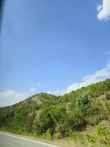 A low-angle shot captures a blue sky with scattered white clouds. In the foreground, a road is visible on the left. The middle ground and background are dominated by a hilly landscape covered in green trees and shrubs. The sun appears to be shining, illuminating the scene. The video is a short, static shot with no discernible action.