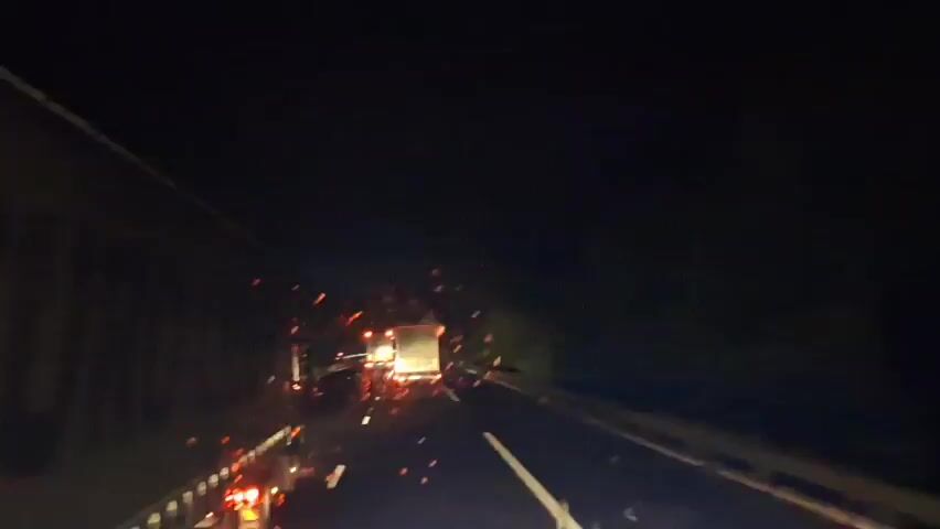 The video shows a nighttime view from inside a vehicle on a highway. Sparks or debris are visible in the air and on the road ahead, suggesting an accident or malfunction. Several large trucks are in front of the vehicle, their taillights illuminated. The road appears wet, and reflections of lights are visible. The overall scene is dark with the exception of vehicle lights and the scattered bright sparks.