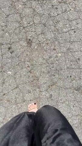 A first-person perspective video shows a person walking on a cracked asphalt surface. The person is wearing black pants that partially obscure their legs and feet. One foot, adorned with nail polish and rings, is visible as the person walks.