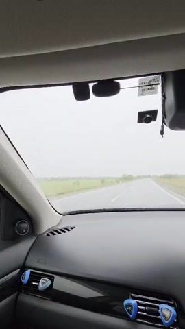 The video captures a first-person perspective from inside a car driving on a straight, two-lane road in overcast or rainy weather, as indicated by the active windshield wipers. The sky is a uniform light grey, and green fields stretch into the distance on both sides of the road, suggesting a rural setting.

Inside the car, the black dashboard is prominent, featuring an infotainment system displaying what appears to be a navigation map alongside media playback controls. The steering wheel, partially visible on the right, is dark with silver accents. Above the dashboard, the rearview mirror is visible, and a dashcam is mounted on the windshield beneath the sun visor. Blue, triangular air vent accents add a pop of color to the otherwise dark interior. A driver's hand is briefly seen on the steering wheel towards the end of the clip. The car's interior appears clean and well-maintained.

Specific text on various objects such as stickers on the sun visor, a small device on the dashboard, and the infotainment screen is too small or blurry to be legibly deciphered. The scene takes place during daylight hours in Бабстово, Russia.