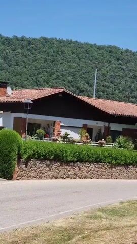 A camera pans across a single-story house with a red tile roof, set against a backdrop of a lush, green, tree-covered hill under a clear blue sky. The house has a dark-colored trim around the roofline and a covered porch area. A low stone wall lined with green bushes and potted plants separates the house from a paved road in the foreground. A lamppost stands near the left side of the house. The overall scene depicts a residential building in a natural, possibly rural, setting.
