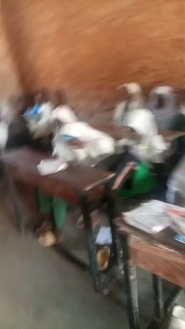 The video shows a classroom. There are students seated at desks. The walls are rough and appear to be made of mud or clay. Most of the students in the front row are wearing white head coverings. There are open books on the desks. The camera pans across the students.