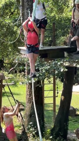 This media file depicts an outdoor adventure course, likely a zipline or ropes course, set within a lush, wooded environment. The scene is brightly lit, suggesting it is daytime with clear weather. In the foreground, a young woman with long hair, wearing a pink and white tie-dye tank top and pink shorts, is reaching upwards, her hands grasping a thick, light blue rope. She appears to be actively participating in an activity.

Above her, on a wooden platform suspended between trees, a woman in a red helmet and red tank top with denim shorts is in motion, holding onto a rope with both hands. She is positioned as if she is either preparing to descend or is in the process of traversing a segment of the course. Behind her, another individual wearing a white t-shirt and dark shorts stands on the same platform, also equipped with safety gear. To the right of the frame, a person in a light brown shirt, dark shorts, and a helmet is visible on another elevated wooden platform, leaning on a railing and observing the activity.

The setting is characterized by mature trees with dense green foliage, a grassy ground below, and visible safety equipment like harnesses and cables. The overall atmosphere suggests an active and recreational event, likely an adventurous outing. The visible text on the white t-shirt of the person behind the woman in the red helmet reads "ROOT." The presence of multiple individuals engaged in this activity implies a group or family outing.