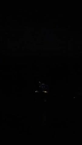 The video captures a nighttime scene with a focus on illuminated objects on the water. The primary subject appears to be a small floating structure or raft adorned with a cluster of colorful lights. These lights, in shades of yellow, red, blue, and green, create a festive and somewhat abstract display against the dark background.

The setting is a body of water, likely a lake or river, given the calm surface that reflects the lights. The time of day is clearly night, indicated by the complete darkness of the sky and the prominence of the artificial lights. There are no visible signs of weather, but the stillness of the water suggests calm conditions.

The activity taking place is not explicitly defined, but the illuminated raft suggests a celebratory event, perhaps a festival or a special occasion. The lights themselves are the main point of visual interest. They are arranged in a somewhat chaotic yet captivating manner, resembling a small, glowing nebula on the water.

There are no visible people or text within the frame. The overall mood is one of mystery and enchantment, brought about by the solitary, brightly lit object in the vast darkness. The reflection of the lights on the water adds another layer of visual depth and a sense of serenity. The context of Batak, Bulgaria, suggests a location in a natural or semi-natural environment where such a display might be set up for a local event.