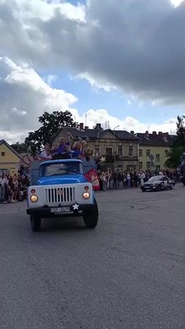 A blue and white truck is driving down a street. A group of children are standing in the back of the truck, waving and smiling. There are people standing on the sidewalk watching the truck go by. The sky is cloudy.