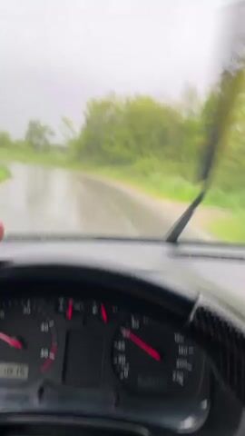 The video shows a first-person perspective of driving a car on a wet road during heavy rain. The windshield wipers are in operation, clearing the rain. The car’s speedometer is visible, as is the black steering wheel. A car is driving towards the camera's car in the opposite direction. The surrounding environment consists of green trees and foliage. The sky is overcast, indicating stormy weather. The car's dashboard is also visible.
