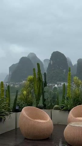 The scene presents an outdoor patio setting under a cloudy sky, with misty mountains visible in the background. Two woven wicker chairs and a round wicker table with a glass top sit on a dark wooden deck, surrounded by large rectangular planters filled with various cacti and succulents. Rain is falling, evidenced by the wet surface of the deck. The mountains in the distance are lush and green, partially obscured by low-hanging clouds or mist.