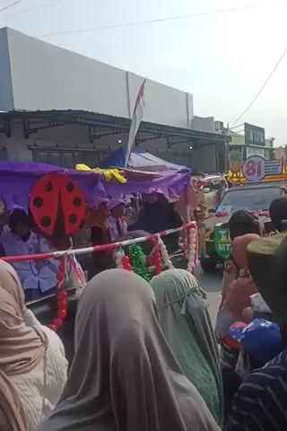 The video shows a parade or procession with several decorated vehicles and a crowd of people. In the foreground, the backs of several people wearing headscarves and hats are visible. In the middle ground, a vehicle is decorated with purple and yellow fabric, and a child in a green outfit is standing on the vehicle. Behind this, another vehicle is adorned with a banner that says "81 INDONESIA MAJU SEJAHTERA DAN MAKMUR" (81 Indonesia Advances Prosperous and Flourishing) and features cartoon pineapples. The vehicle also has a sign that says "TK MTA". Indonesian flags are visible in the background, along with buildings and tents that suggest a street fair or event. The crowd appears to be watching the procession. The weather seems to be sunny.