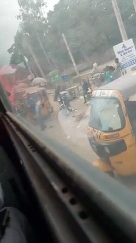 The video, viewed from inside a vehicle through a smudged window, shows an outdoor scene with several auto-rickshaws, commonly known as tuk-tuks, parked on a dirt road. A line of these yellow vehicles is visible, some with passengers or drivers inside. In the foreground, a man in a blue shirt and grey pants is interacting with a tuk-tuk driver. Further back, a person on a motorcycle is observed, and other individuals are scattered around the area, some appearing to be vendors or bystanders. A sign for "Mini Resort & Garden" is visible on a pole. Trees line the background, and the sky is overcast. The overall impression is a bustling roadside environment, possibly a transportation hub or market area.
