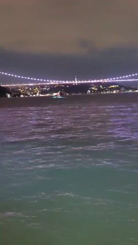 The video shows a body of water with waves at night. In the background, a bridge is lit up with many lights, and a city skyline is visible. The sky is dark and cloudy. The water in the foreground reflects the lights from the bridge and the city, giving it a purple and green hue.
