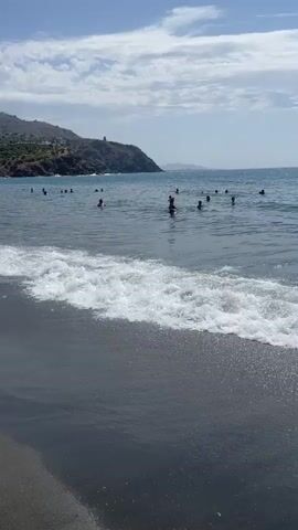 The video shows a crowded beach on a sunny day. The camera pans from the water's edge to reveal people swimming and playing in the ocean, with buildings and hills in the background. The beach itself appears to be sandy and dark in color. The water is clear and blue, with small waves breaking on the shore. Beach umbrellas are set up along the sand, and there are many people relaxing under them. The overall atmosphere appears to be one of leisure and enjoyment.