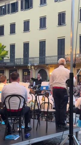A brass band plays outdoors on a raised stage in front of a light yellow building with dark shutters. A conductor, wearing a black shirt, directs the band with his left hand raised. Several musicians, all wearing white shirts and dark pants with red belts, are seated on chairs facing the conductor. Some musicians are holding instruments, including a tuba and a trombone. An Italian flag waves on the right side of the frame. Spectators are seated in the background, facing the stage. The scene appears to be in a town square or courtyard.