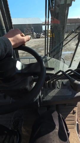 The video shows a person operating a forklift, likely in a lumberyard or construction site. The camera is mounted inside the forklift cab, providing a first-person view. The operator's left hand is on the steering wheel. The forklift's controls, levers, and pedals are visible. Through the windshield, stacks of lumber and puddles on a dirt ground are visible. The sky is clear and blue, suggesting a sunny day. The forklift appears to be stationary. A large warehouse or building is visible in the background.