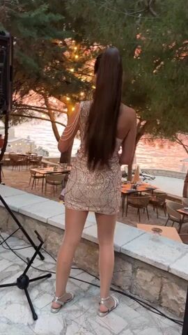 This media file captures a woman playing the saxophone on a stone patio. She is facing away from the camera, with her long dark hair pulled into a high ponytail. She is wearing a sparkly, sequined, strapless dress and silver sandals. The saxophone is gold-colored and she holds it in playing position. 

The setting appears to be an outdoor dining area, with tables and chairs visible in the background. Trees with string lights are scattered throughout the scene, casting a warm glow. The sun is setting over the water, creating a colorful sky in shades of orange and pink, indicating it is dusk. The overall atmosphere seems festive and relaxed. The woman's posture and the act of playing the saxophone suggest she is performing.
