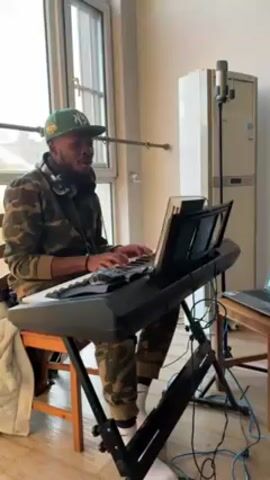 A man is sitting and playing a digital keyboard, while singing. The man is African American, with a beard, and he is wearing a green baseball cap, camouflage jacket, and pants, with white socks and headphones around his neck. He sits on a brown wooden chair. The keyboard is black, on a black stand, and has sheet music in front of it. To the right of the keyboard is a wooden table with a laptop on it and a white cable hanging down. Further to the right is a tall, white appliance with a microphone in front of it. To the left is a large window with sunlight coming through.