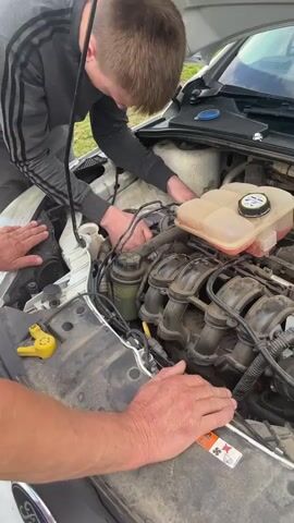 Two people are working on the engine of a white car with the hood open. One person, wearing a gray hoodie, is leaning into the engine bay and appears to be touching something near the front of the engine. Another person's hands, presumably an adult, are resting on the fender and engine cover, guiding or assisting. The engine is visible with its manifold and various components. The scene appears to be outdoors, possibly a driveway or yard, with green grass and some other vehicles in the background. The lighting suggests it is daytime.