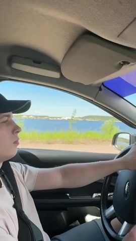 A young man wearing a black baseball cap and a tan t-shirt is driving a car. He is wearing a seatbelt. The car is moving along a road next to a large body of water, possibly a lake or river. There are trees and bushes along the shoreline, and the sky is blue with some clouds. The sun is shining. The interior of the car is visible, including the dashboard and the man's arm resting on the steering wheel. The view outside the window is a landscape.