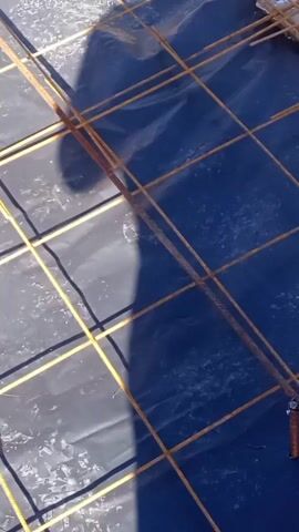 A man in a green hooded jacket and blue sweatpants with the Puma logo stands outside and strokes his chin while looking at a wire rebar grid laid over a black tarp. The view then shows two men kneeling on the wire grid, one using a wooden level. A plastic chair and tablecloth can be seen in the background.