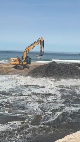 An excavator is positioned on a sandy beach next to a body of dark, foamy water. The excavator, a yellow Caterpillar model, is actively scooping dark material from a large pile on the beach and depositing it into the water, causing significant disturbance and churning. The water appears dark and agitated, with white foam and debris visible on the surface. In the background, the ocean stretches to the horizon under a clear blue sky. There are faint outlines of boats or ships in the distance on the ocean. The overall scene depicts an active construction or environmental cleanup operation at the edge of the water.