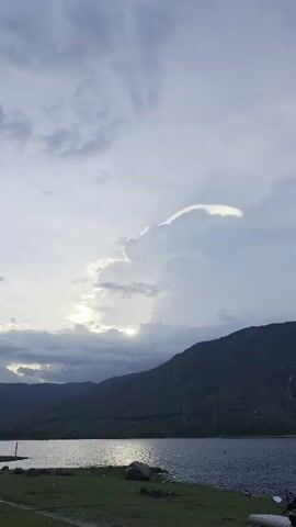 The video shows a scenic view of a lake surrounded by mountains under a cloudy sky. The sun is obscured by clouds, creating a diffused light. Notably, there is a cloud with a bright, iridescent edge, possibly due to iridescence or circumhorizontal arc. The camera zooms in to focus on this cloud formation. The landscape is visible in the foreground, with some vegetation along the lake shore. A voice in the background can be heard saying words relating to photography.
