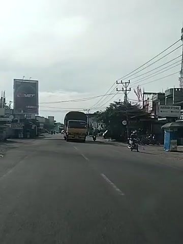 The video opens with a wide shot of a two-lane road. A large yellow truck with a tarp-covered trailer is driving towards the camera in the left lane. On the right side of the road, several small shops and a cell tower are visible. A motorcycle is seen passing the truck on the right side of the road. As the camera moves closer to the truck, the video focuses on its right side, showing the driver's side of the cab and the trailer. The truck then passes out of frame. The video concludes with a shot of the road ahead, showing more shops and parked vehicles. The sky is overcast, suggesting it may be daytime. The road appears to be in a semi-urban or rural area.