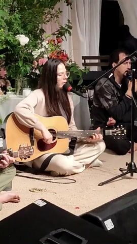 A medium shot shows a woman playing an acoustic guitar and singing into a microphone while sitting on the floor. She has long dark hair and is wearing glasses and a light-colored sweater and pants. Another person is visible to her right, holding a microphone. The background includes floral decorations and stage lighting.