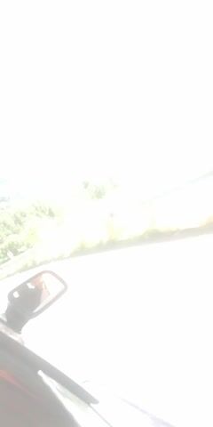 A video from inside a vehicle shows a scenic mountain landscape with greenery and a bright blue sky. The vehicle is driving on a road with a guardrail. The driver's side mirror reflects the driver's face and arm. The camera is tilted, giving an unusual perspective of the scene.