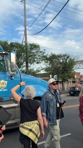 A group of about 20 people are gathered in a parking lot or street near a large, bright blue semi-truck, some of whom are taking photos or videos of the vehicle. The truck has what appears to be a stylized wheat stalk or floral design on the side. The people appear to be a mix of ages, and some are wearing masks. The scene is outdoors and the weather appears to be mild. There are utility poles and wires overhead, and other vehicles and buildings are visible in the background.