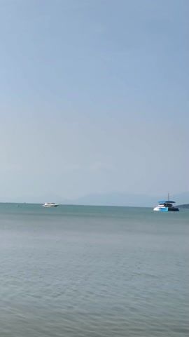 The video shows a daytime view of a calm sea. Initially, a blue and white catamaran and a white boat can be seen in the distance. As the video progresses, an orange boat moves across the frame towards the right. In the background, there is an island or landmass with hills and buildings. The video concludes with a wider view of the sea, revealing several sailboats and a building with a tower on the distant shore. The sky is clear and blue. At one point, a person's hand briefly enters the frame at the bottom of the screen.