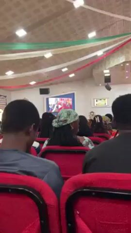 The video shows an audience sitting in red chairs, likely in a church or auditorium setting. The focus is on the back of the heads of the attendees. A screen displays a person speaking, likely the pastor or a speaker, with pink and blue lighting on the stage. There are decorative streamers hung from the ceiling. The camera zooms in slightly on a woman in the audience wearing a green and brown patterned headscarf. The overall atmosphere suggests a service or presentation.