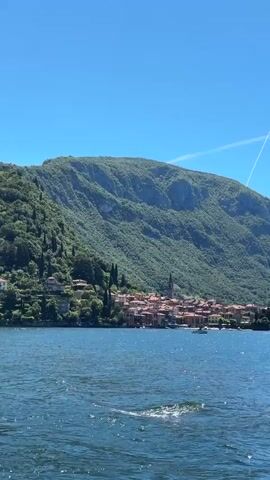 A wide shot captures a view of a village nestled at the base of a lush, green mountain range overlooking a body of water. The water is a deep blue and appears to have gentle waves. The village is composed of buildings with red-tiled roofs, and a church steeple is visible. The mountain is covered in dense trees and vegetation. The sky is a clear, bright blue with a faint contrail from an airplane. A small white boat is visible on the water in the middle distance, moving from left to right. The overall impression is a scenic, tranquil landscape.