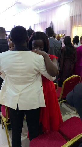 A man and a woman are hugging in a room full of people. The woman is wearing a red dress, and the man is wearing a white jacket and black pants. They are standing among rows of chairs. Other people are milling around. A stage is visible in the distance. A sign says “ANCHORED.”
