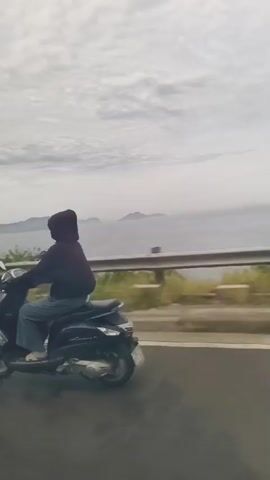The video shows a person riding a dark-colored scooter on a road. The person is wearing a dark hooded jacket and blue jeans, with their face obscured by the hood. The scooter is moving forward, and the rider's left hand is on the handlebars. The road appears to be asphalt with a yellow line on the left side. In the background, there is a metal guardrail, vegetation, and a body of water with hills or mountains in the distance. The sky is cloudy. The camera is positioned to the side and slightly behind the scooter, capturing its movement along the road.