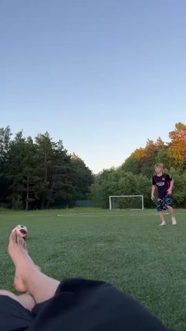The video begins with a close-up of a person's bare feet and legs, lying on grass. A soccer ball is visible near the feet. The camera then pans up to reveal a young boy in a soccer jersey and shorts running on a grassy field towards the right side of the frame. In the background, a soccer goal is visible, surrounded by trees. The sky is clear and blue. The boy kicks the ball, which then travels across the field. The camera shifts perspective, showing the boy lying on the grass. Another shot shows a bird flying in the sky. The video ends with a shot of the boy still lying on the grass.