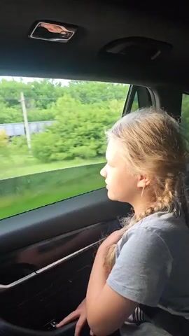 A young girl with braided hair looks out the window of a moving car. Outside, trees pass by with a train in the distance. The car's interior is visible, showing the door panel, handle, and the ceiling light.
