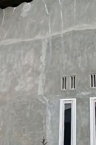This media file captures a natural scene from Meunasahkumbang, Indonesia, during what appears to be a heavy downpour. The primary focus is on a building's exterior wall, which has a rough, textured, grey finish. High up on the wall, three small rectangular windows are visible, each with vertical slats. Below these, two larger windows, framed in white, occupy the lower portion of the scene.

In front of the building, a small, dark-leaved plant with a bushy top stands prominently. To its left, a pile of reddish-brown bricks is stacked. The ground in the foreground is muddy and wet, with visible raindrops creating ripples and splashes on the surface, indicating active rainfall. Small patches of green grass are scattered across the ground. The overall impression is one of a quiet, natural event unfolding in a residential or semi-rural setting. The weather is clearly wet, with rain falling heavily enough to create significant puddles and splashes. There is no visible text in the media file. The time of day is difficult to ascertain due to the overcast conditions likely caused by the rain, but it appears to be daytime. The mood is calm and serene, despite the intensity of the rain.