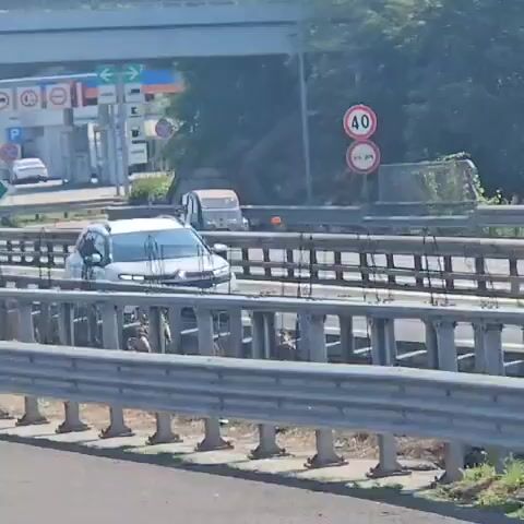 The video pans across a highway overpass. In the background, there is a gas station with several cars and a white van. A small, light blue car is driving on the highway. A large truck is visible in the distance. There are also guardrails and trees.