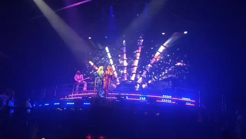 A stage is lit up with blue lights and two women are singing on it. The women are wearing colorful, sparkly outfits. Behind them is a large screen displaying a colorful, concentric design. Two other musicians, one playing guitar and one playing a keyboard, are also on stage. A spotlight shines down on the performers. An audience is visible in the foreground.
