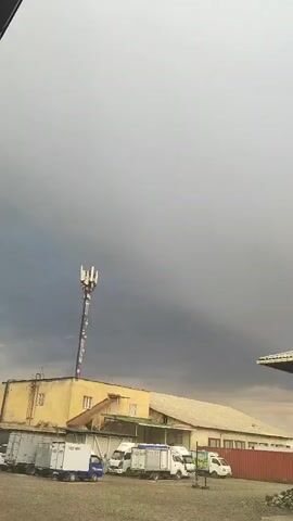 The video shows an exterior view of a commercial or industrial area under a dark, overcast sky. In the foreground, a gravel lot contains several white delivery trucks parked in a row. Behind the trucks, a long, light-colored building with a white roof stretches across the frame. To the left of the building, a yellow structure with a cell tower on top stands out. The sky is filled with heavy, gray clouds, suggesting impending rain or a storm. The overall atmosphere is somber and muted.