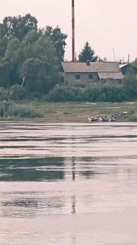 The video captures a landscape scene from the perspective of a body of water. In the foreground, the water's surface is rippled and reflects the overcast sky. In the middle ground, a bank of land is visible, with a stretch of green vegetation and some scattered debris or objects. Several small boats or rafts are moored near the bank. In the background, there are several structures. On the left side, a building with a tall, red-and-white striped chimney stands among trees. To the right of that, a cluster of houses is nestled within a wooded area, with a dirt road leading up to one of them. Further to the right, industrial elements become apparent, including what appears to be a crane or construction equipment, along with more buildings and trees. The overall impression is of a semi-rural or industrial area situated alongside a river or large body of water. The sky is uniformly pale, indicating a cloudy or hazy day.